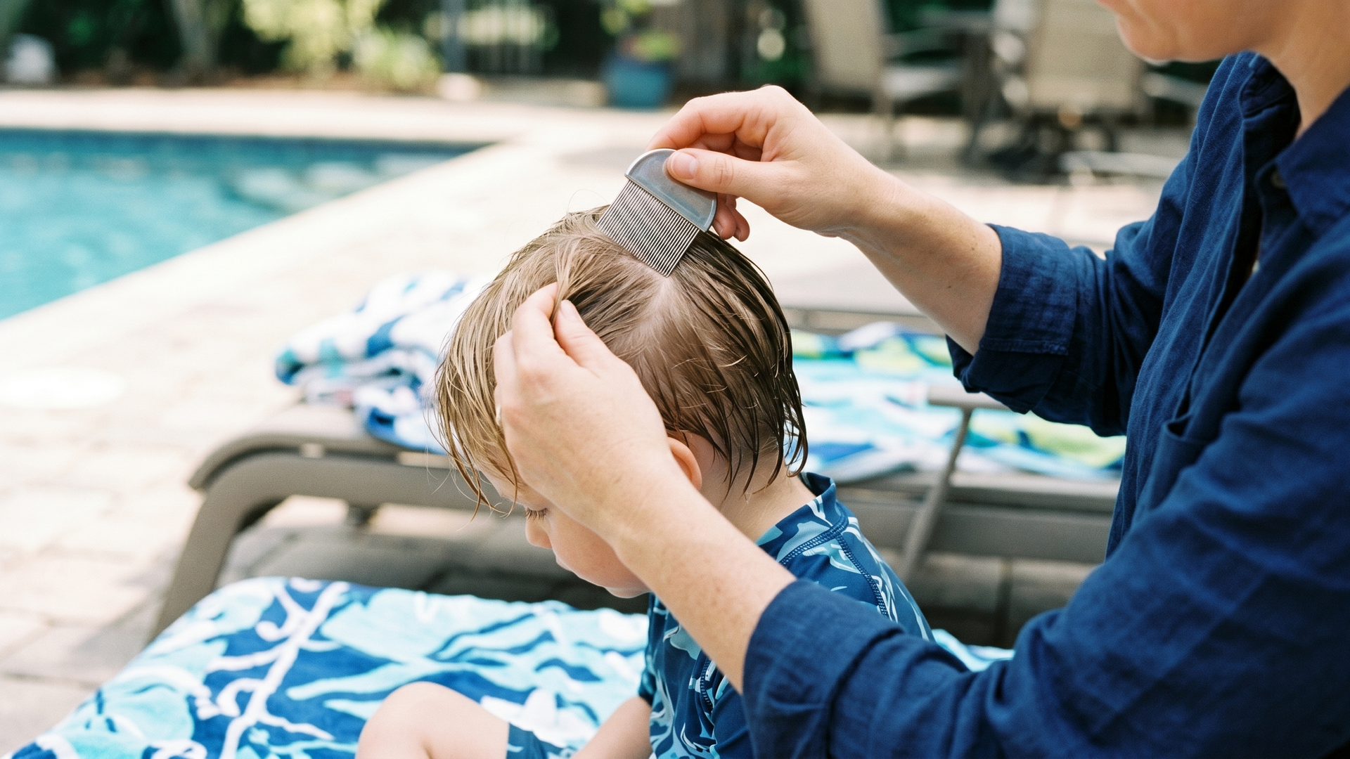 Parent doing a fine-toothed lice comb check on a child sitting on a poolside towel before a swim day