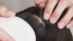 Mother in a soft cream long-sleeve standing at a brightly lit bathroom mirror gently pinching a tiny white speck between her thumb and forefinger above her young daughter's parted brown hair, with a stainless-steel fine-tooth metal nit comb and a folded white hand towel resting on the counter - illustrating the at-home texture test the article walks parents through.
