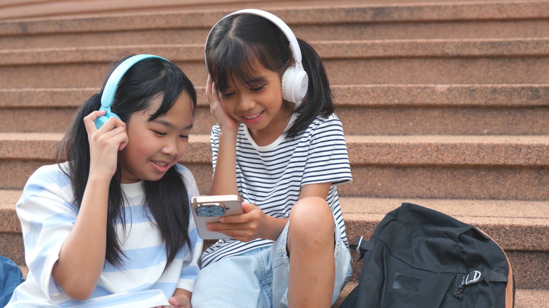 Two school-age girls sharing headphones while sitting together.