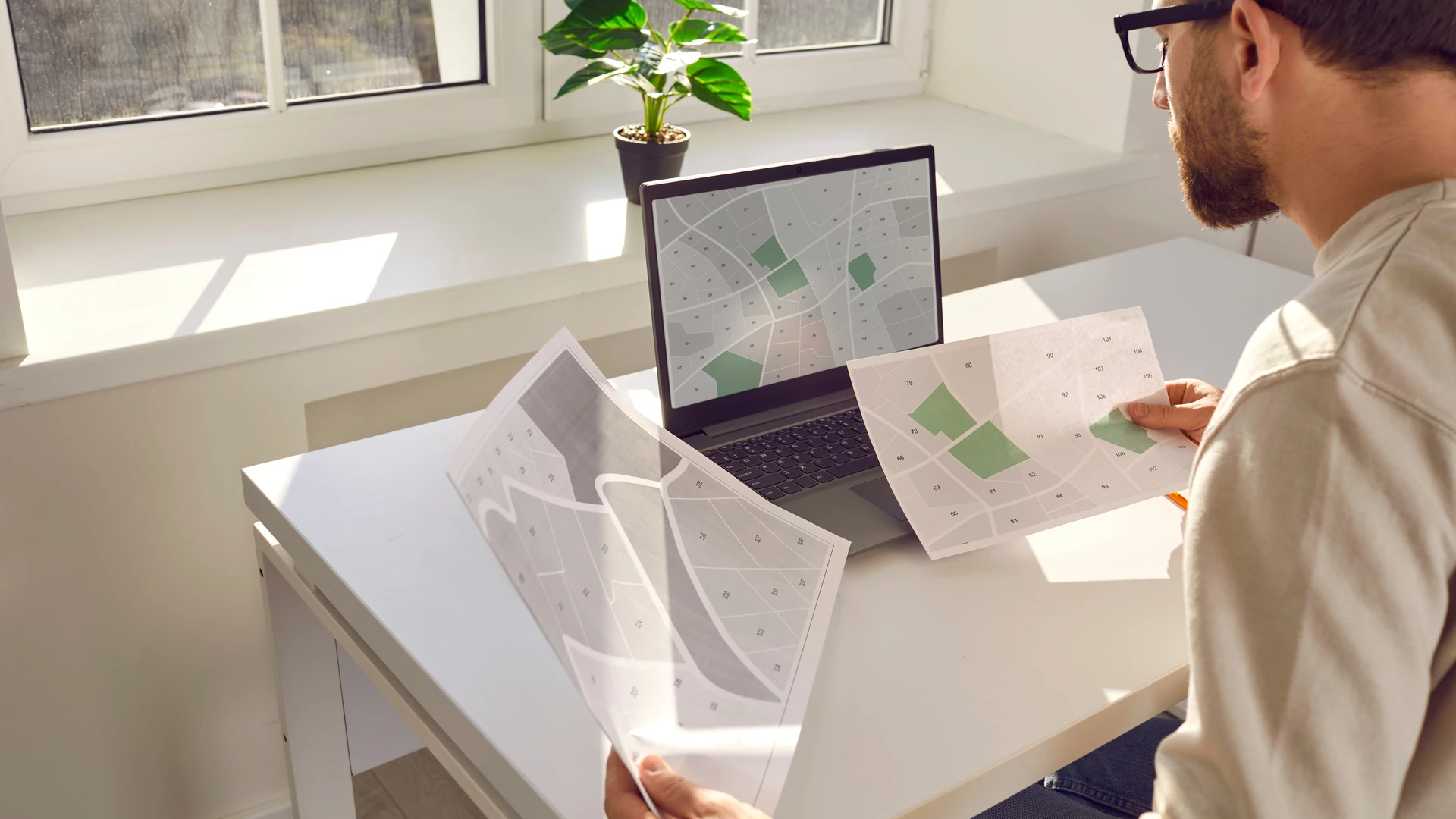 A man at a desk comparing printed maps with shaded parcels against a map on his laptop