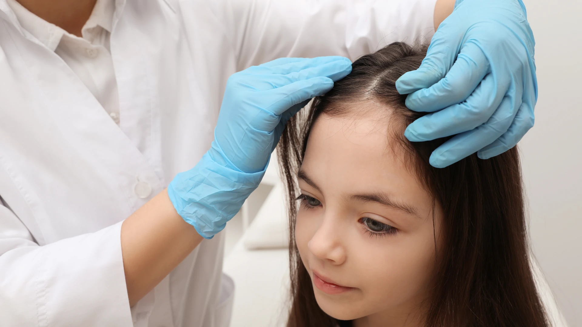 Gloved hands part a calm young girl's brown hair to check her scalp for signs of lice