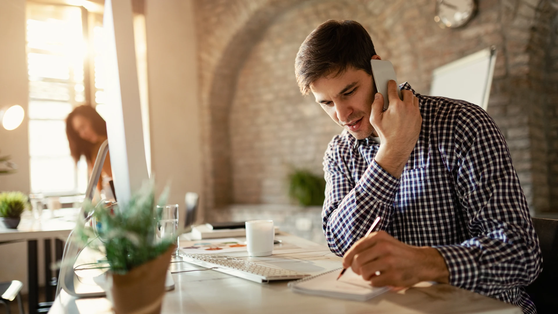Man at an office desk taking notes in a notebook while talking on a phone call