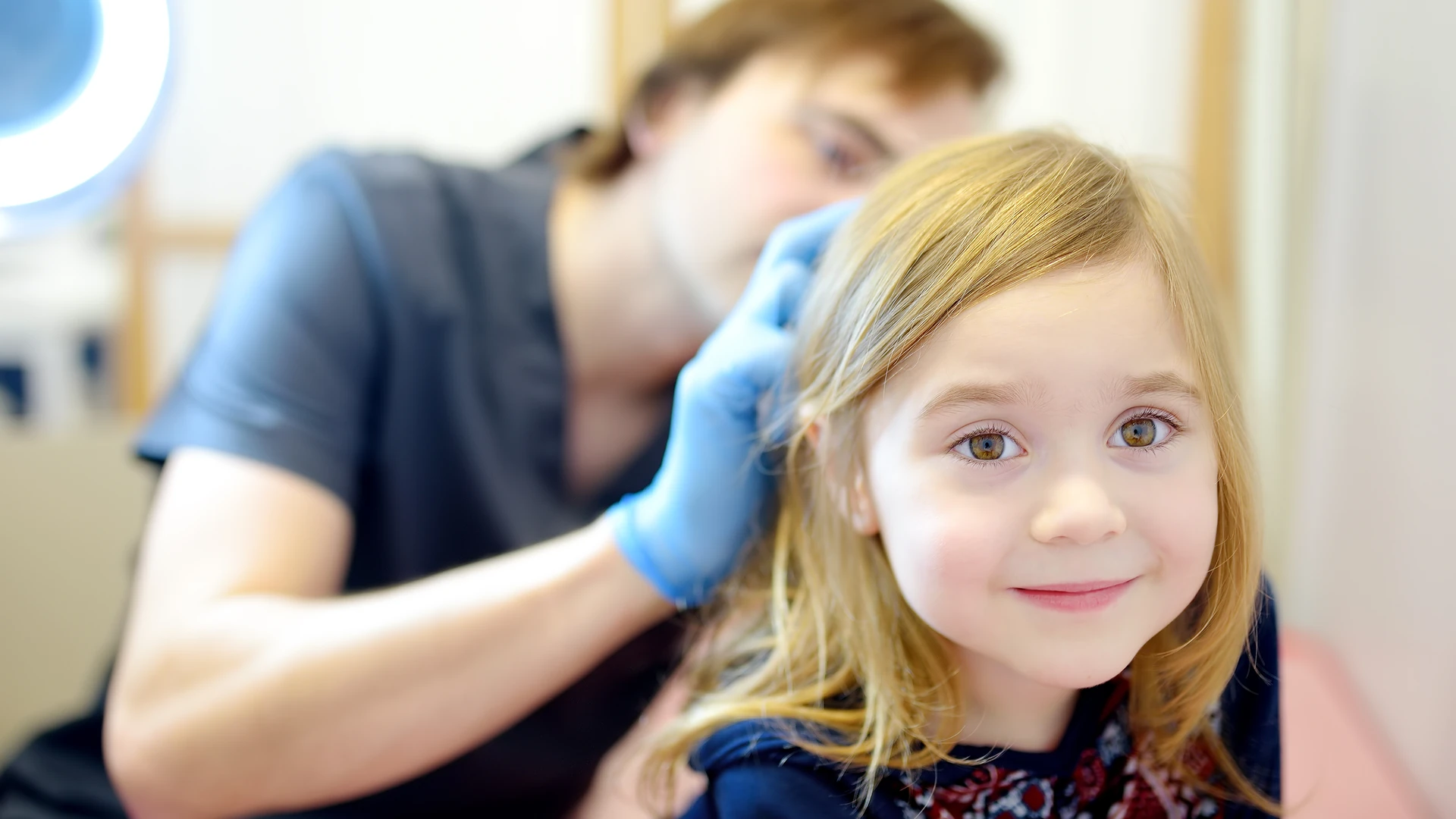 Smiling girl sits calmly while a gloved professional checks her long blonde hair for lice